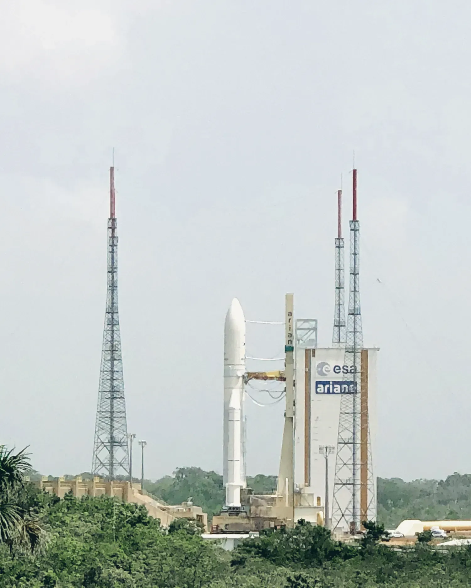 Ariane 5 vue de loin, blanche, dressée sur son pas de tir entre de hauts pylônes paratonnerres, à côté d’un portique aux logos de l’ESA et d’Ariane. La forêt au premier plan, sous un ciel voilé.
