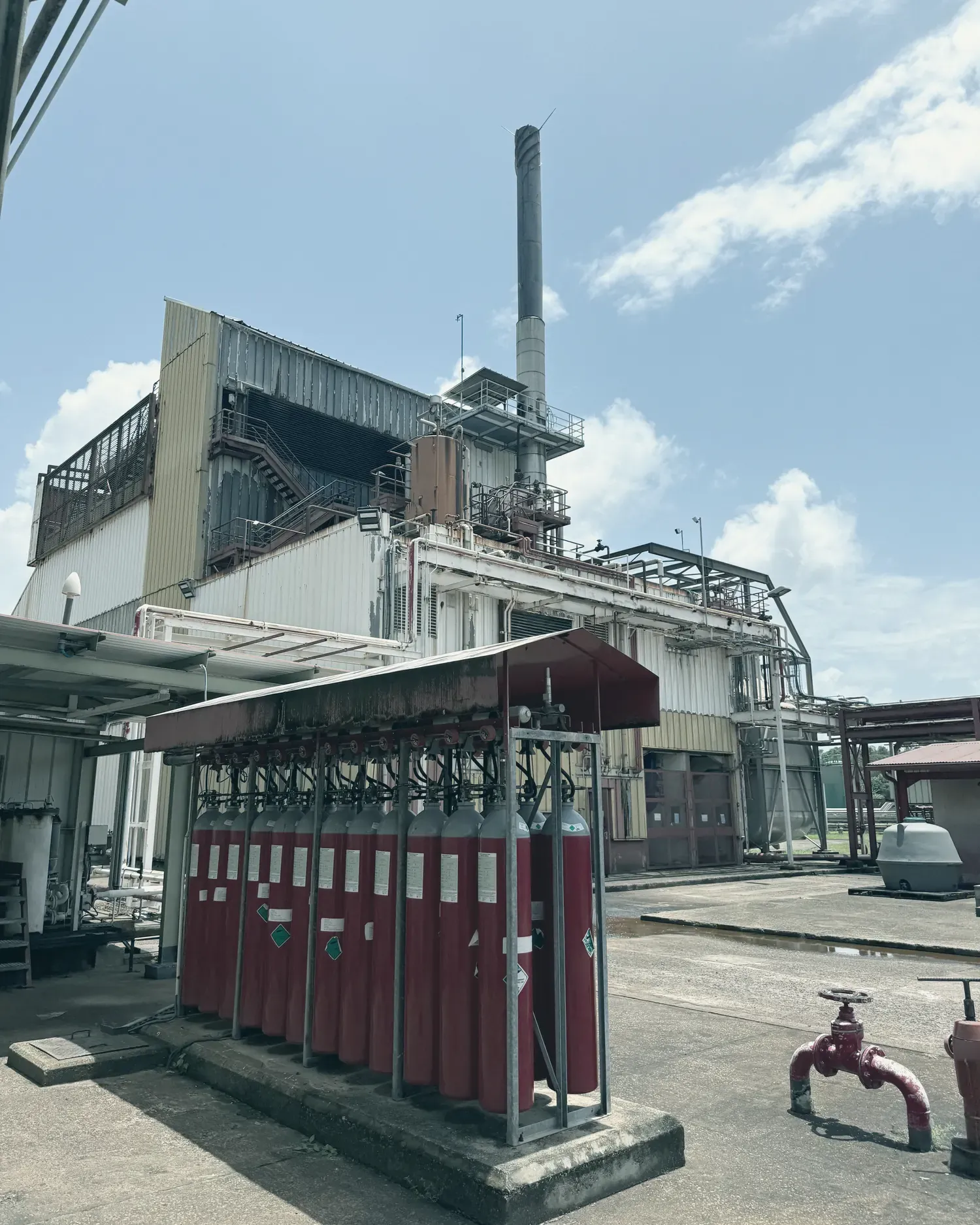 Une rangée de bouteilles de gaz rouges sous un auvent, devant le bâtiment d’une centrale thermique et sa cheminée.
