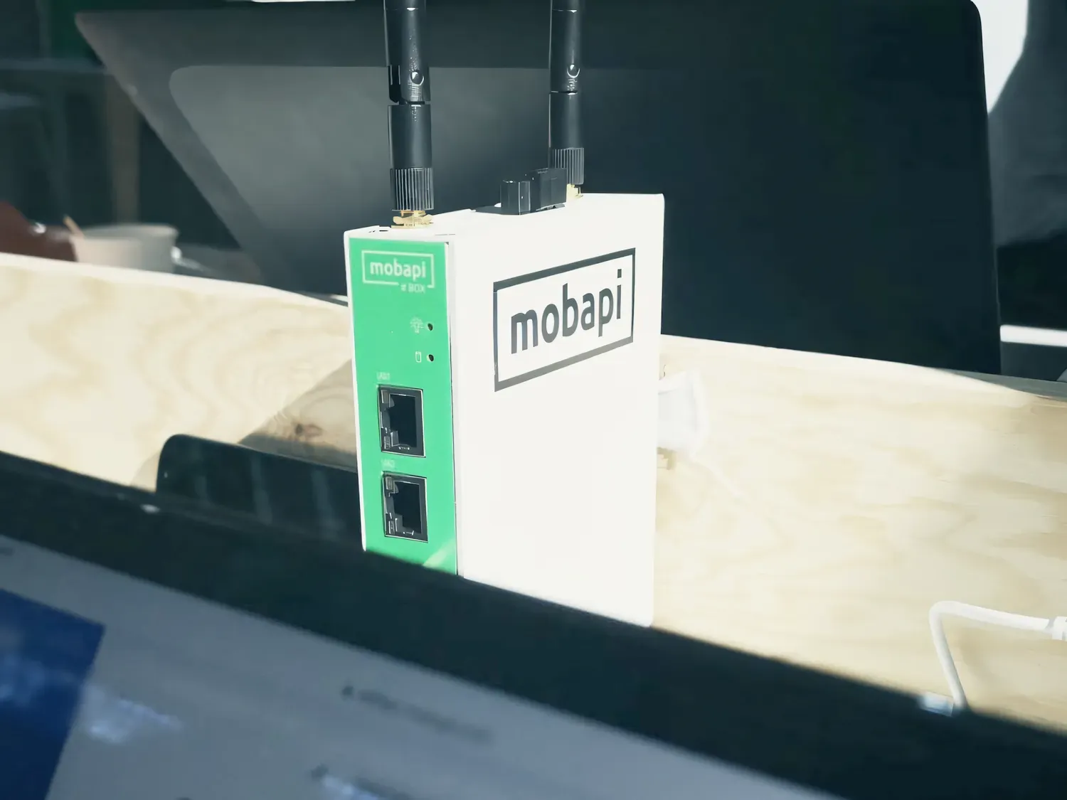 La Mobapi.BOX debout sur une table en bois : un boîtier blanc et vert au logo Mobapi, deux antennes et deux prises réseau.