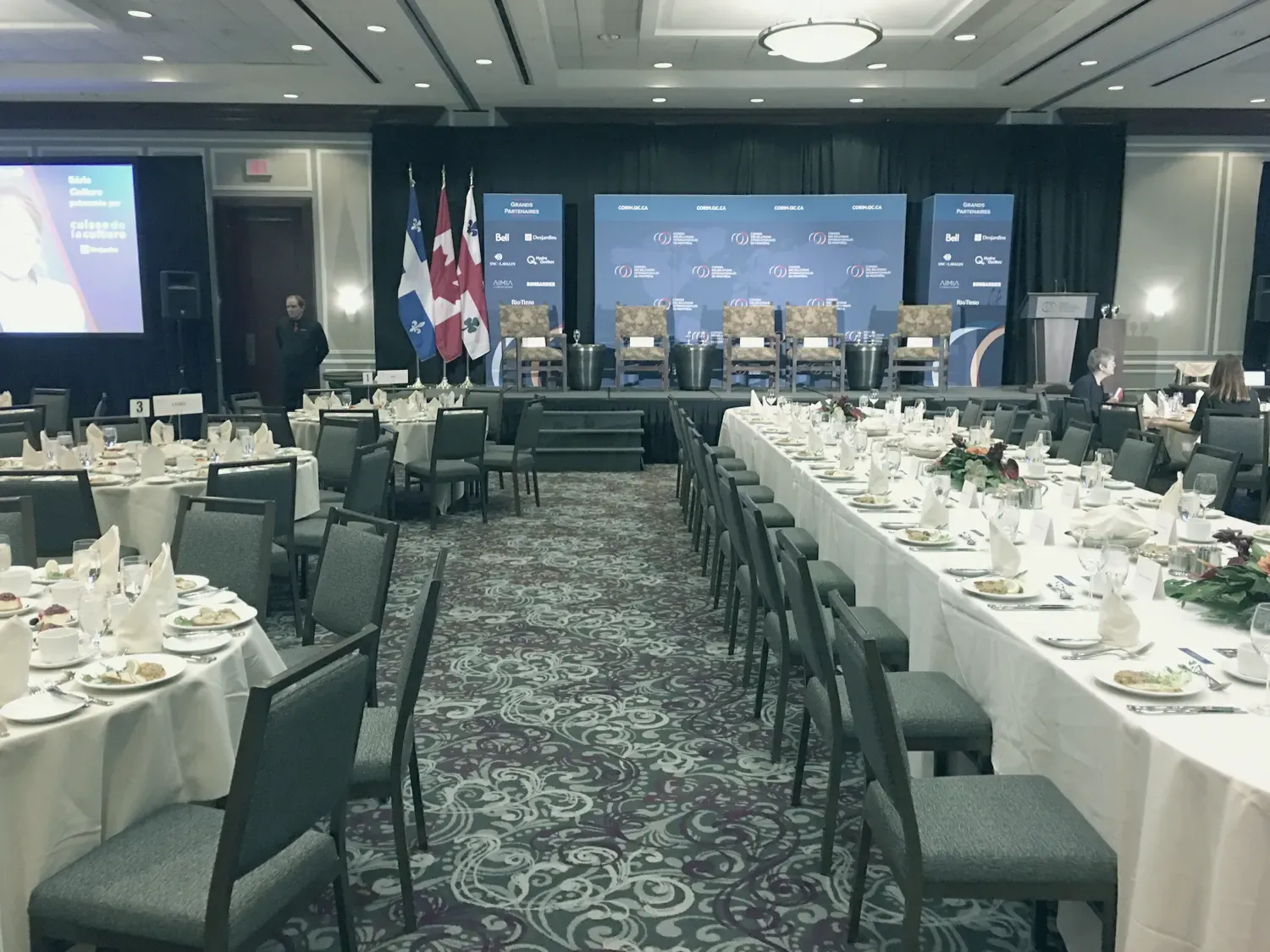 Une salle de déjeuner-conférence aux tables dressées, face à une estrade décorée des drapeaux du Québec, du Canada et de Montréal.