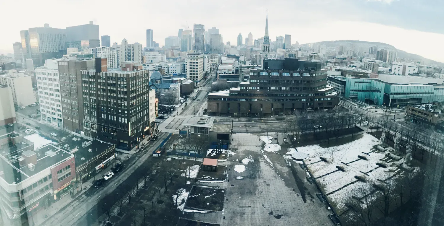 Le centre-ville de Montréal vu d’un étage élevé : des tours, un clocher, le mont Royal au fond et, au premier plan, une place où il reste de la neige.
