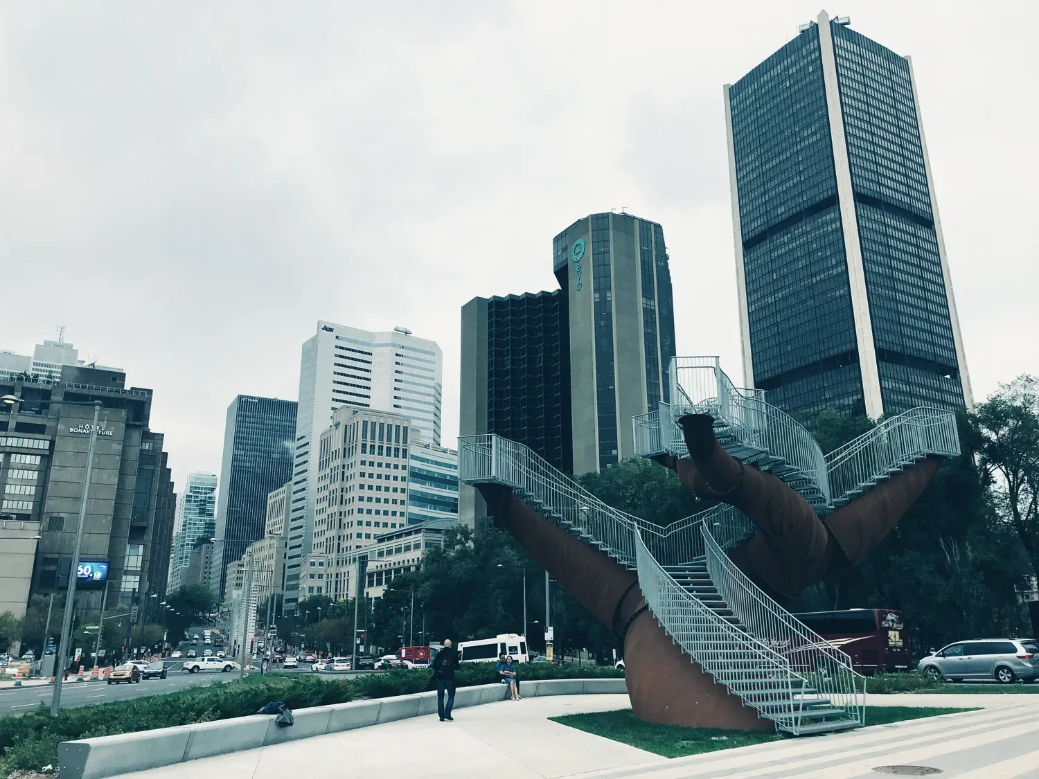 Une sculpture monumentale en acier couleur rouille, faite de deux escaliers enlacés, devant les tours du centre-ville de Montréal, sous un ciel gris.