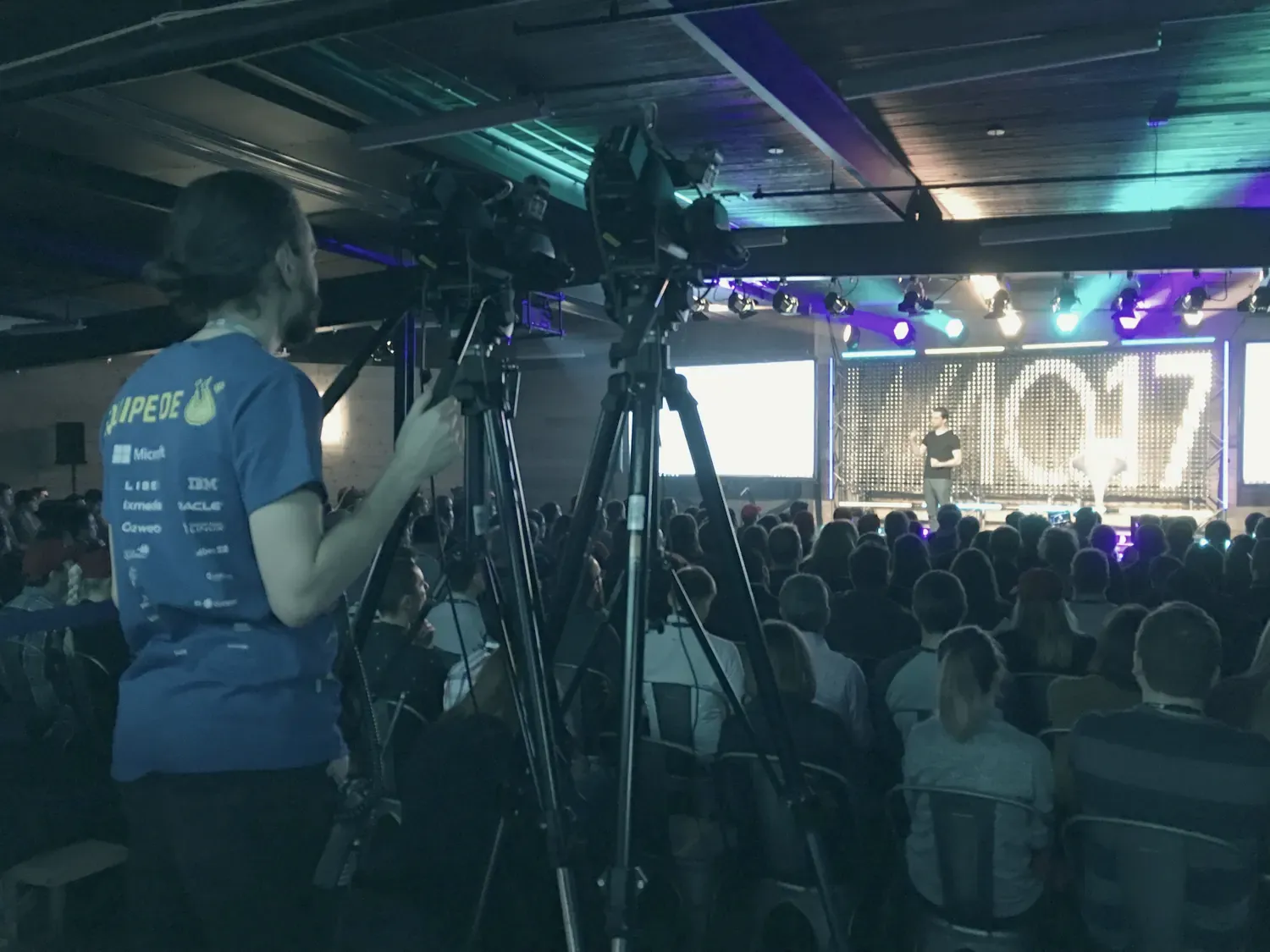 Une salle de conférence plongée dans le noir : le public de dos, un cadreur et ses caméras au premier plan, un orateur sur une scène éclairée.