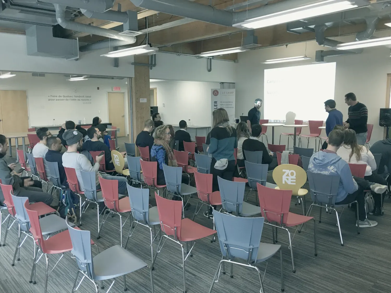 Une salle aux chaises rouges et grises : une vingtaine de personnes, assises ou debout, devant un écran de projection.