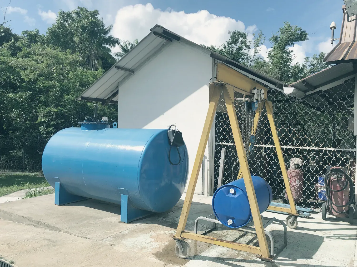 Une cuve de stockage bleue, posée sur une dalle à côté d’un portique de levage jaune qui tient un fût, devant un local technique.