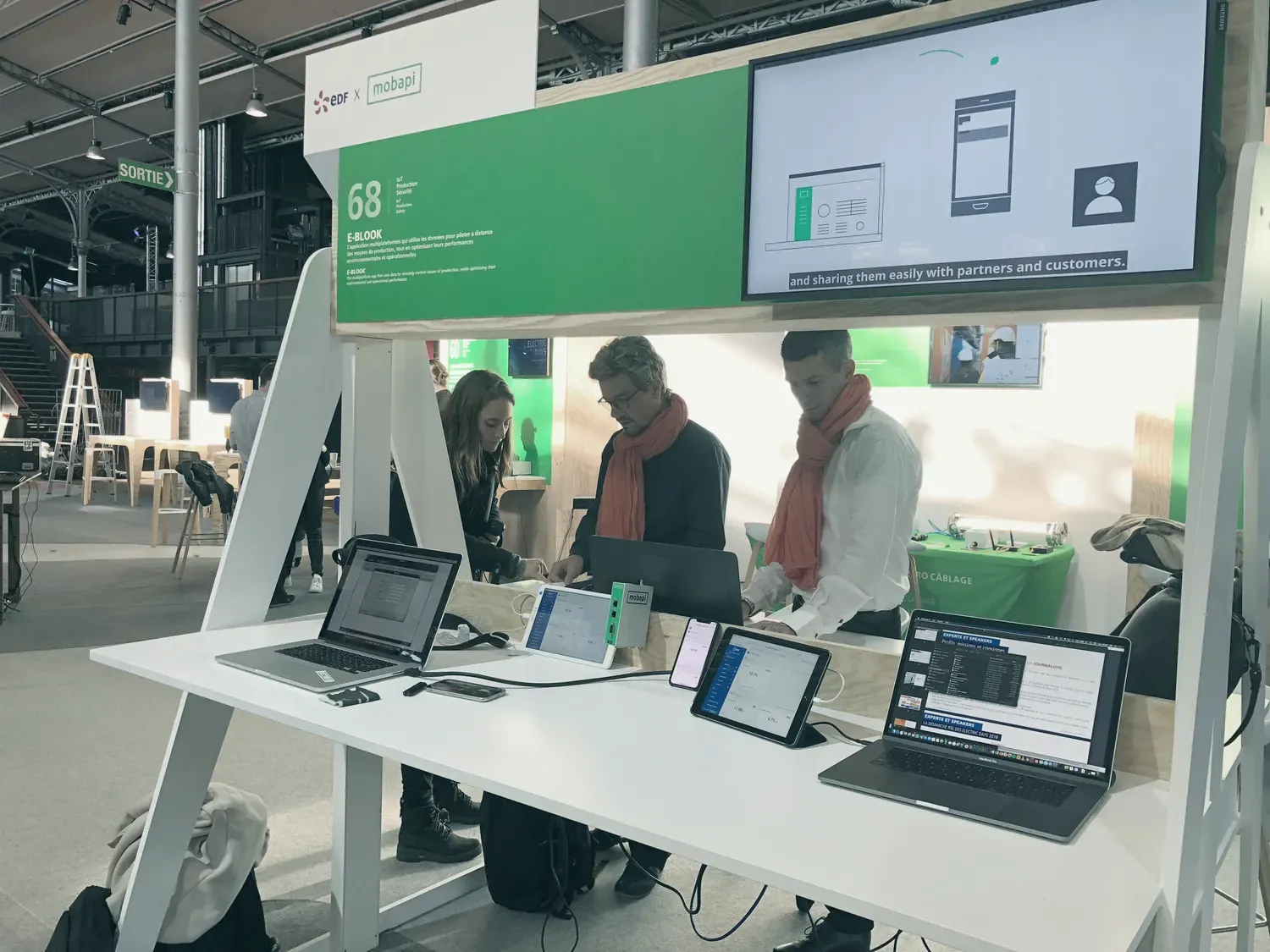Le stand « EDF x mobapi » sous une halle à charpente métallique : trois personnes derrière une table blanche où sont posés des ordinateurs, des tablettes et une Mobapi.BOX verte, sous un écran de présentation.