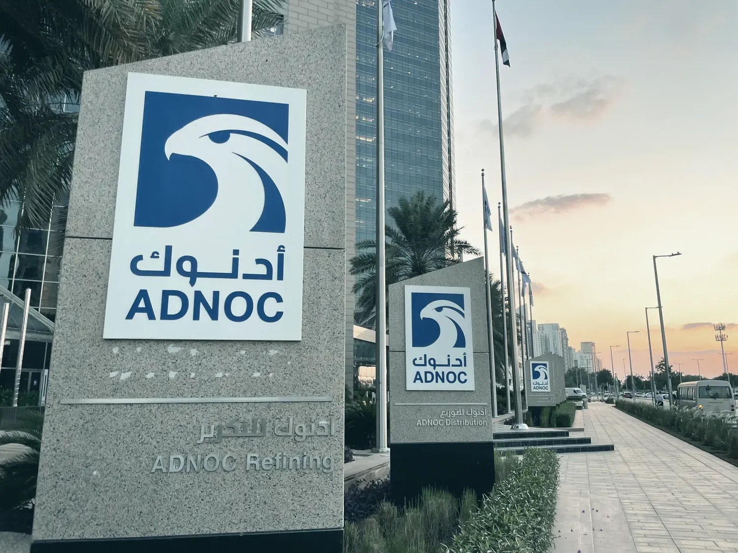 Les enseignes d’ADNOC et d’ADNOC Refining devant un immeuble de bureaux, le long d’une avenue bordée de palmiers.