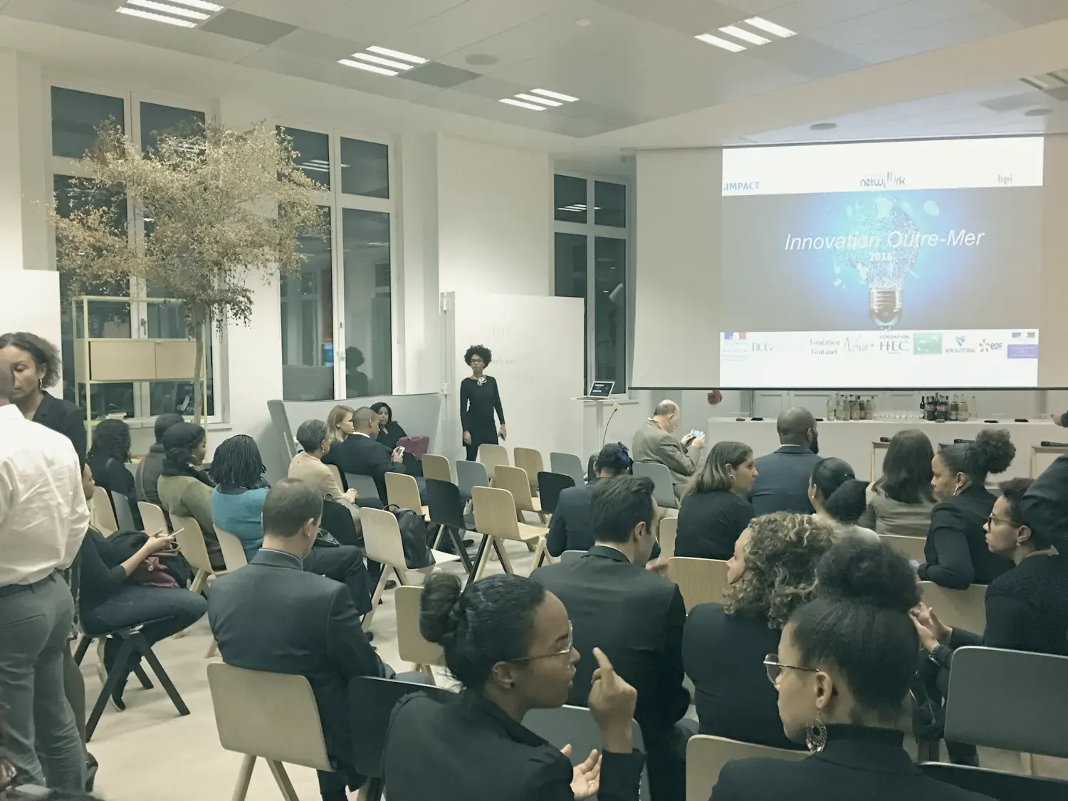 Une salle pleine, vue du fond : le public assis de dos, une oratrice debout, et un grand écran qui affiche « Innovation Outre-Mer 2016 » au-dessus des logos des partenaires.