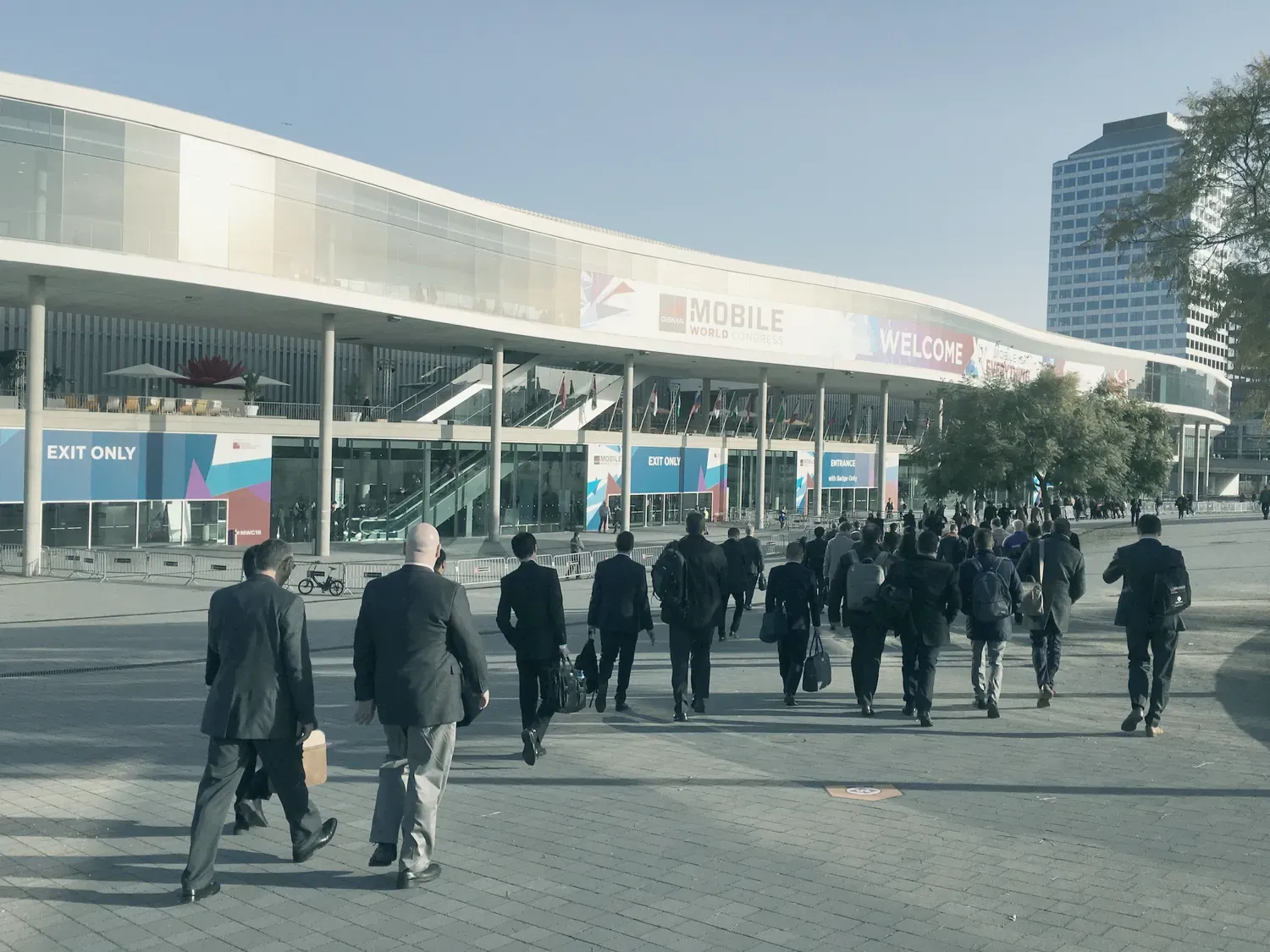 Des visiteurs en costume, vus de dos, marchent vers l’entrée du salon : une longue façade blanche qui porte la bannière « Mobile World Congress » et le mot « Welcome ».