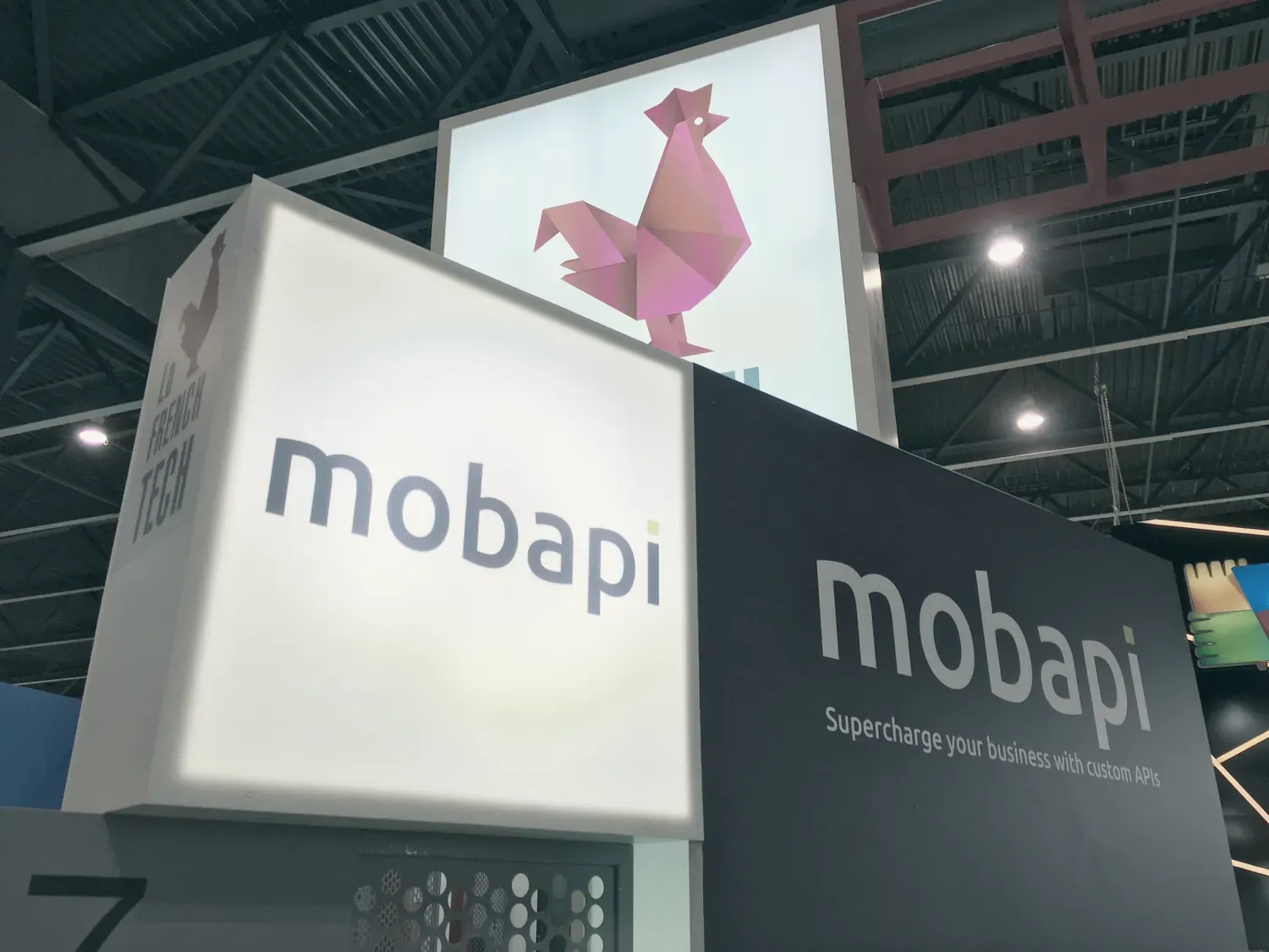 Deux enseignes « mobapi », l’une lumineuse, l’autre sur fond noir avec le slogan « Supercharge your business with custom APIs », sous le coq rouge de la French Tech.
