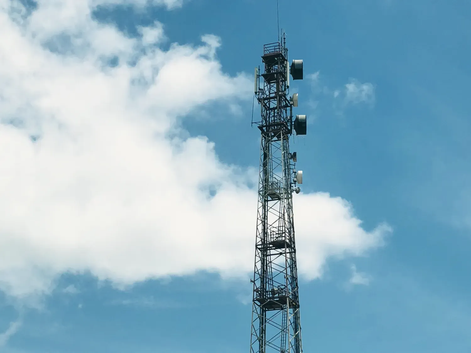 Un pylône de téléphonie mobile en treillis, chargé d’antennes et de paraboles, sur un ciel bleu traversé de nuages.