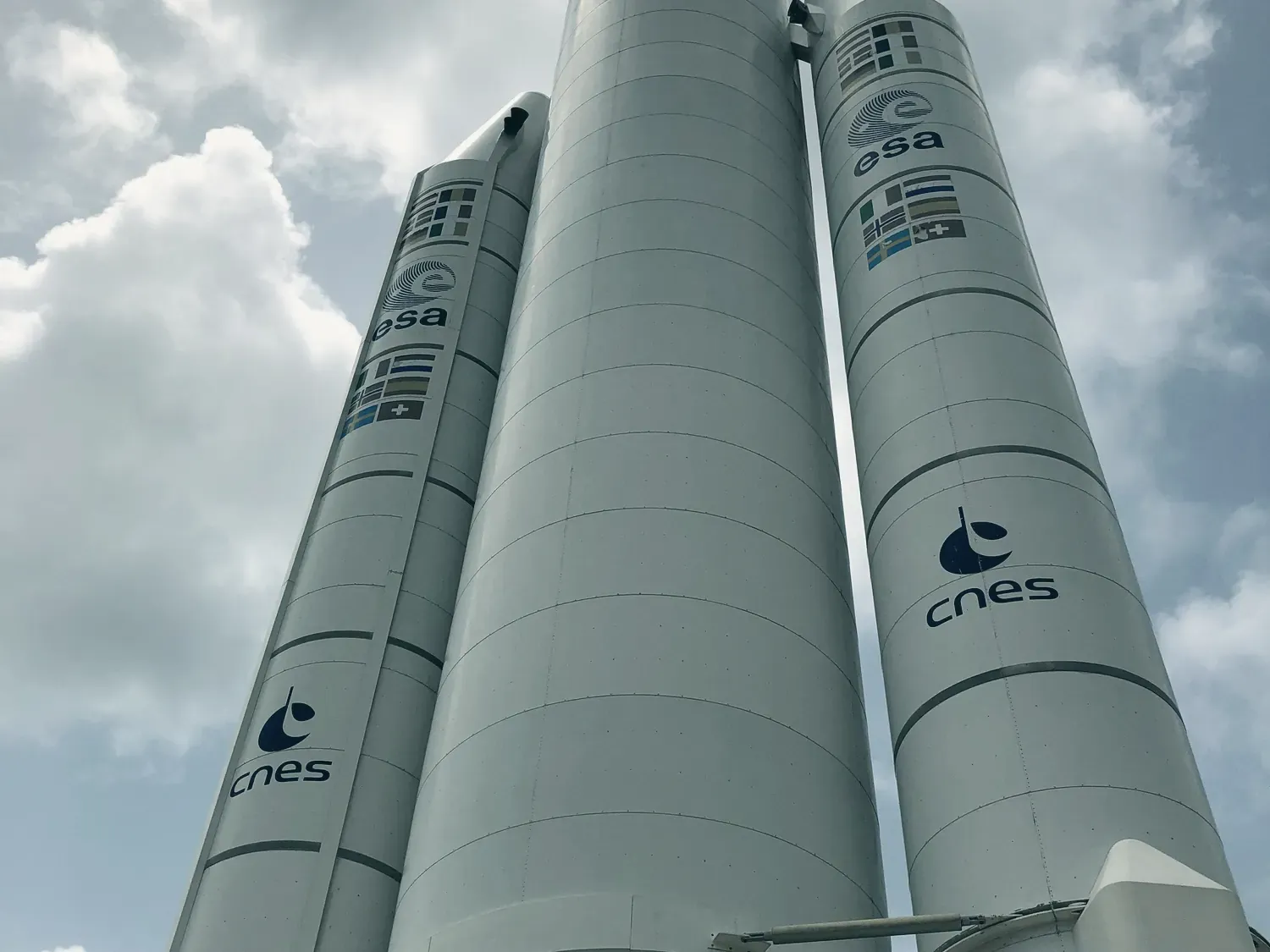 Ariane 5 vue du pied, en contre-plongée : le corps central et deux propulseurs blancs aux logos de l’ESA et du CNES, sous un ciel nuageux.