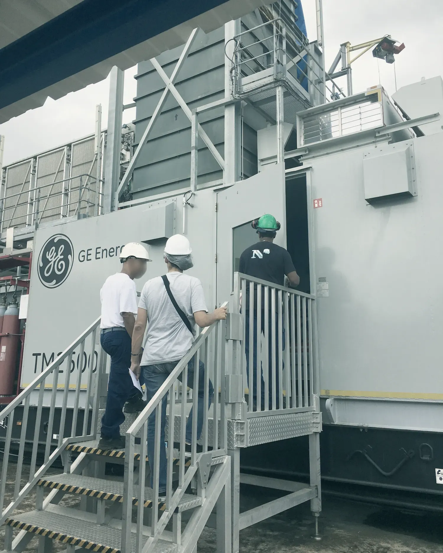 Trois techniciens montent l’escalier d’accès d’une turbine aérodérivée GE TM2500, sur une centrale thermique.