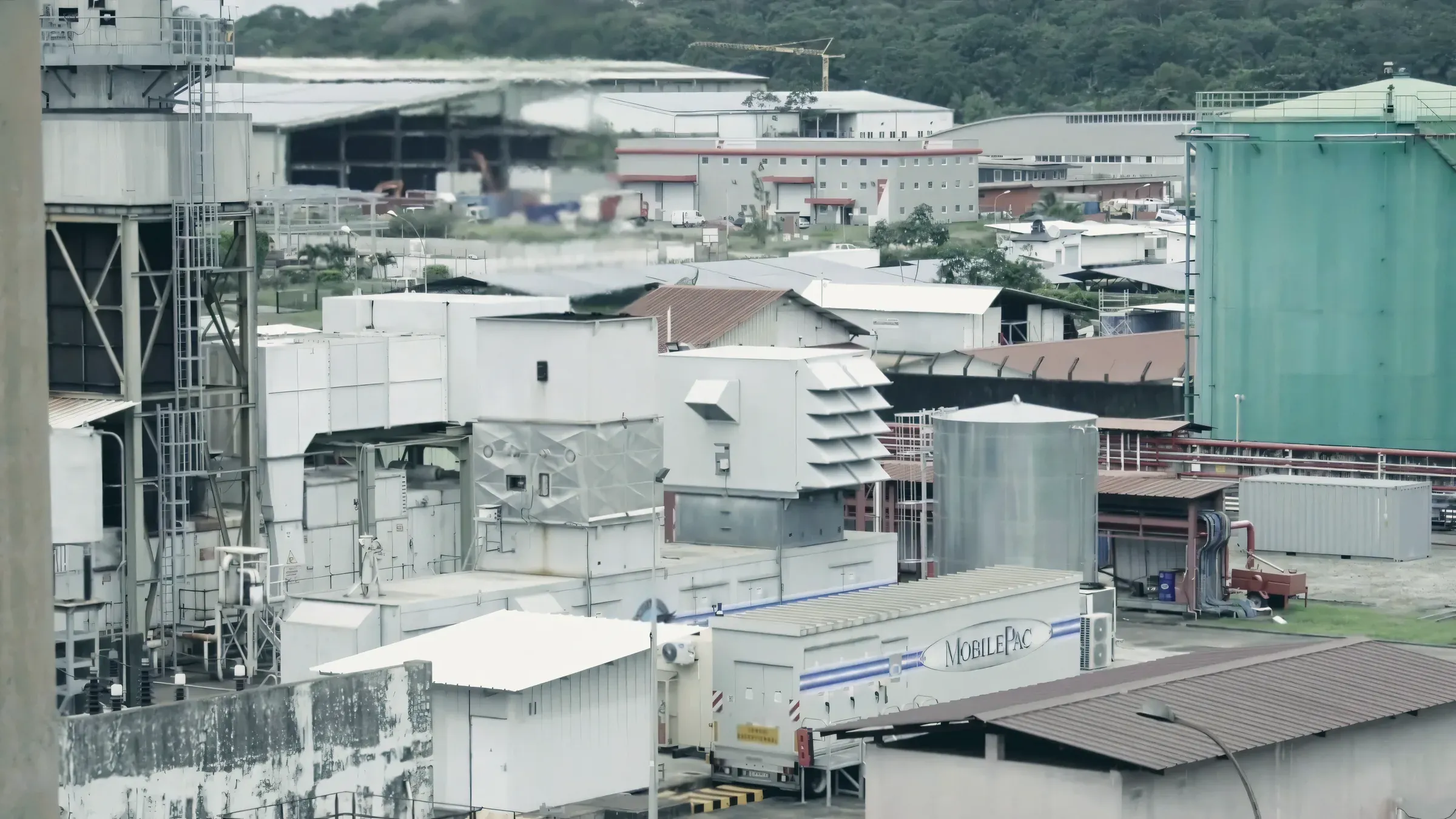 Une turbine à combustion sur remorque et ses caissons, à côté d’une cuve métallique et d’un grand réservoir vert, devant des bâtiments industriels et une colline boisée.