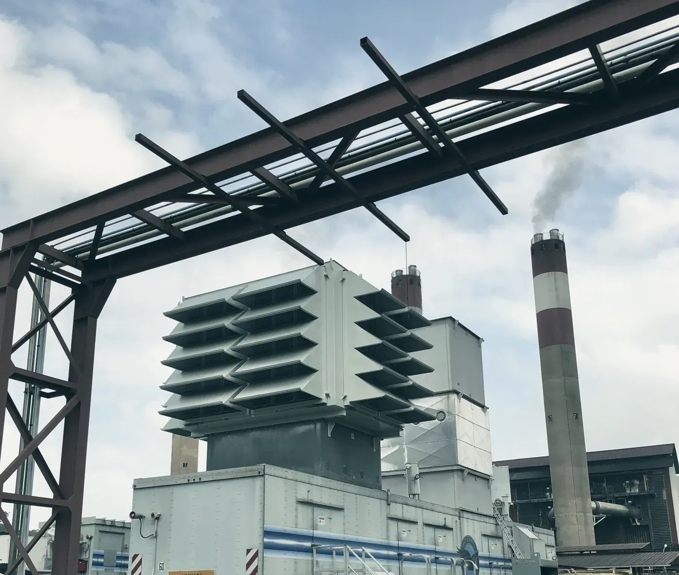 Le caisson d’entrée d’air d’une turbine à combustion, sous un portique de tuyauteries, avec deux cheminées en arrière-plan.